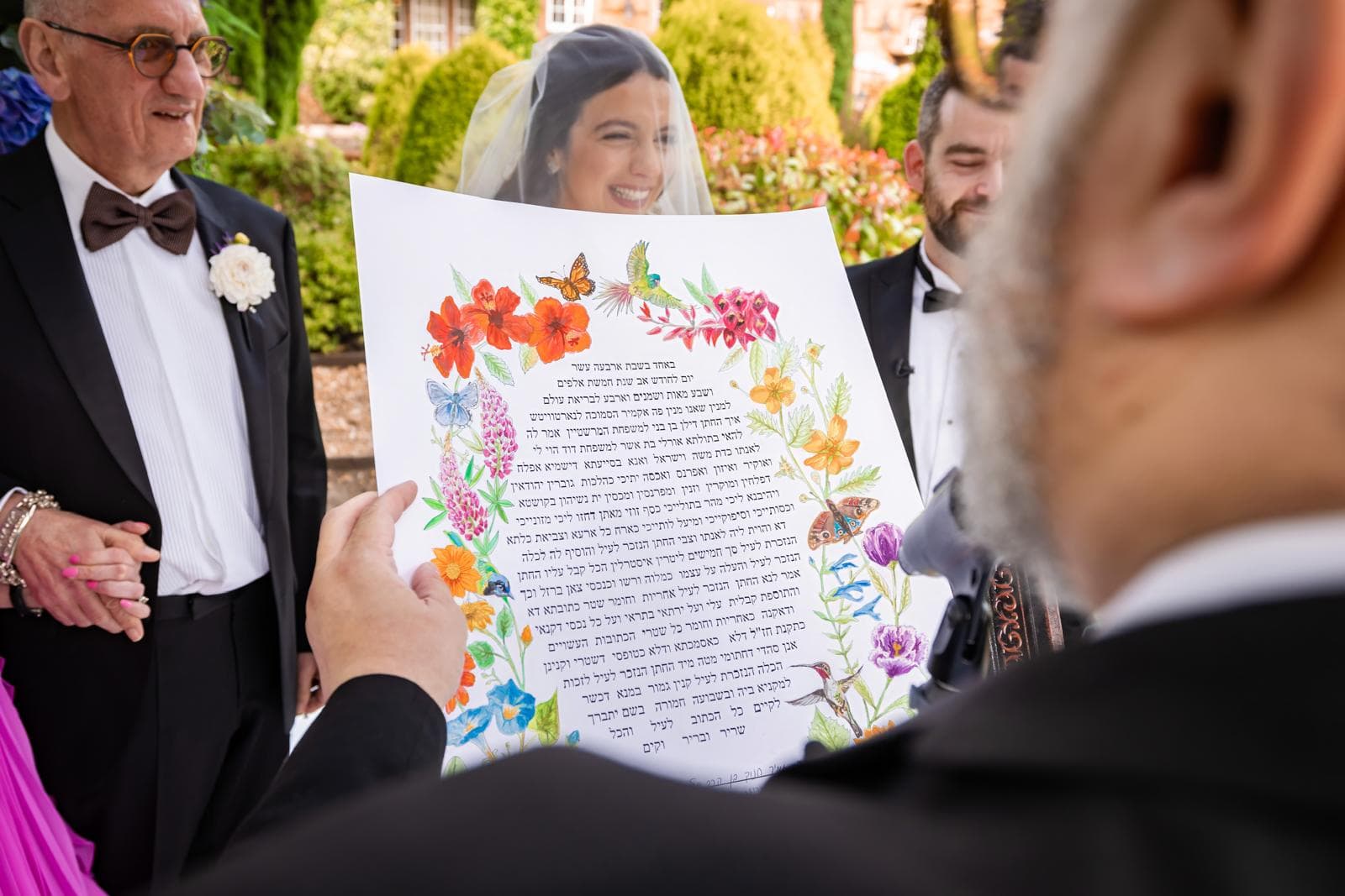 A colourful floral ketubah being read at a Jewish wedding, with the couple smiling behind it